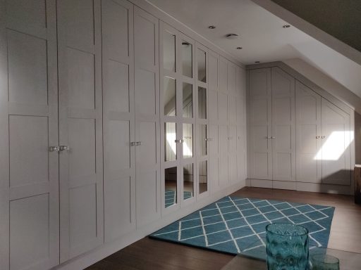 Cashmere Grey wardrobe with frame and panel doors. Light, airy attic room with white cabinets, a large mirror, and a turquoise patterned rug.
