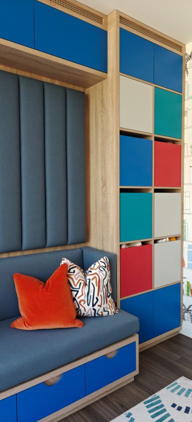 Playroom seating with lower drawers and floor to ceiling storage. Brightly coloured storage unit beside a cozy blue bench with decorative cushions.