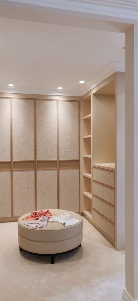 Sand Beige dressing room. Wallpapered doors with integrated handles Modern room with built-in shelving and a circular ottoman in a neutral colour scheme.