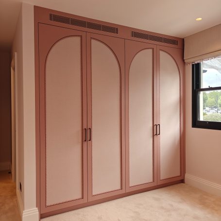 Antique rose finish wardrobes featuring elegant arched panel detailing, integrated top ventilation grilles, bobble bead details. Modern four-door wardrobe with a pink and arched design in a light room.